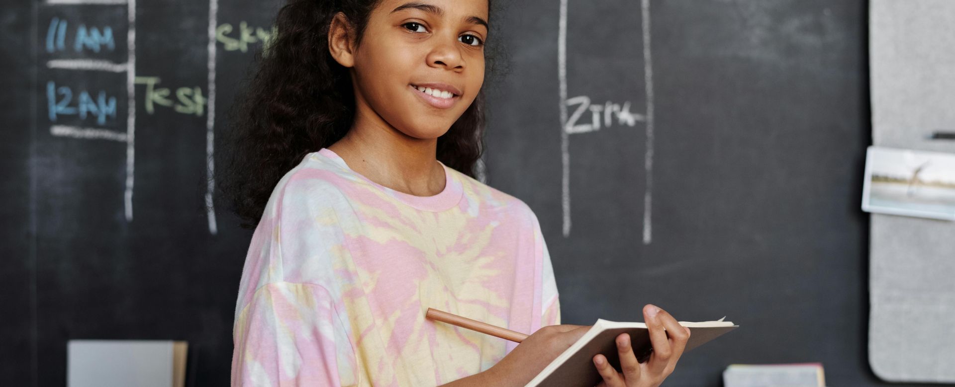 Teenage girl smiling while holding a notebook in a classroom setting with a chalkboard behind her.