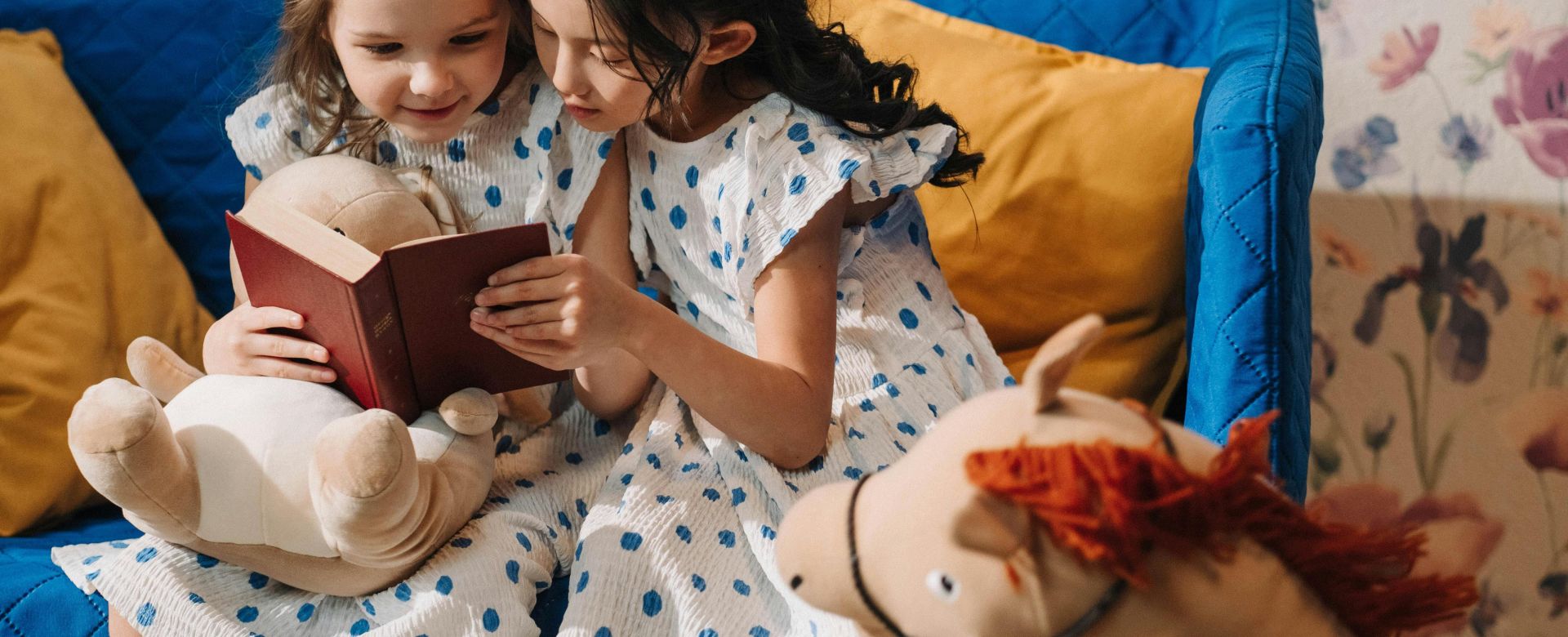 Two young girls in polka dot dresses happily reading together on a cozy sofa with stuffed toys.